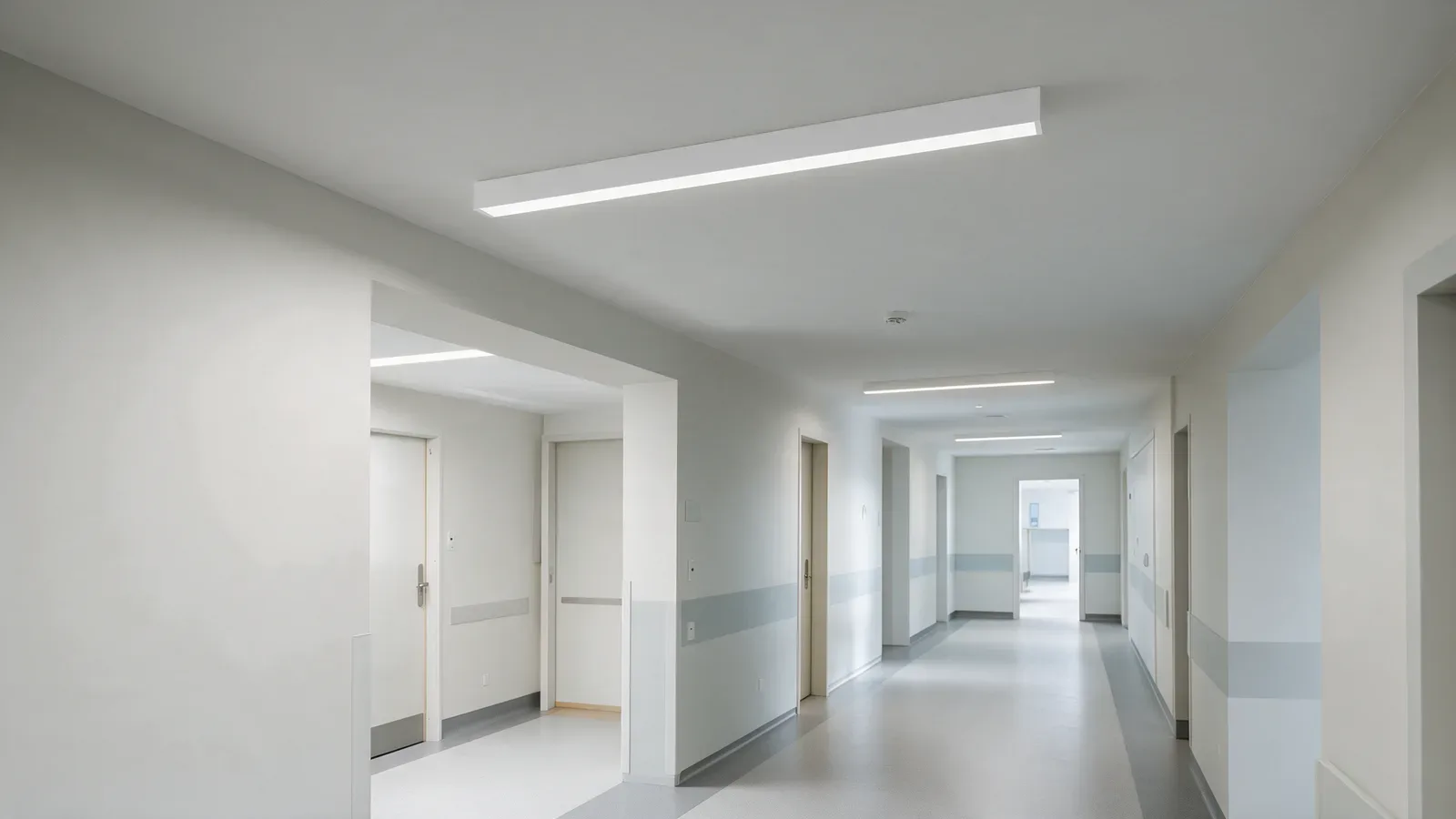 Pasillo público de hospital, despejado, con luminarias lineales blancas de superficie.