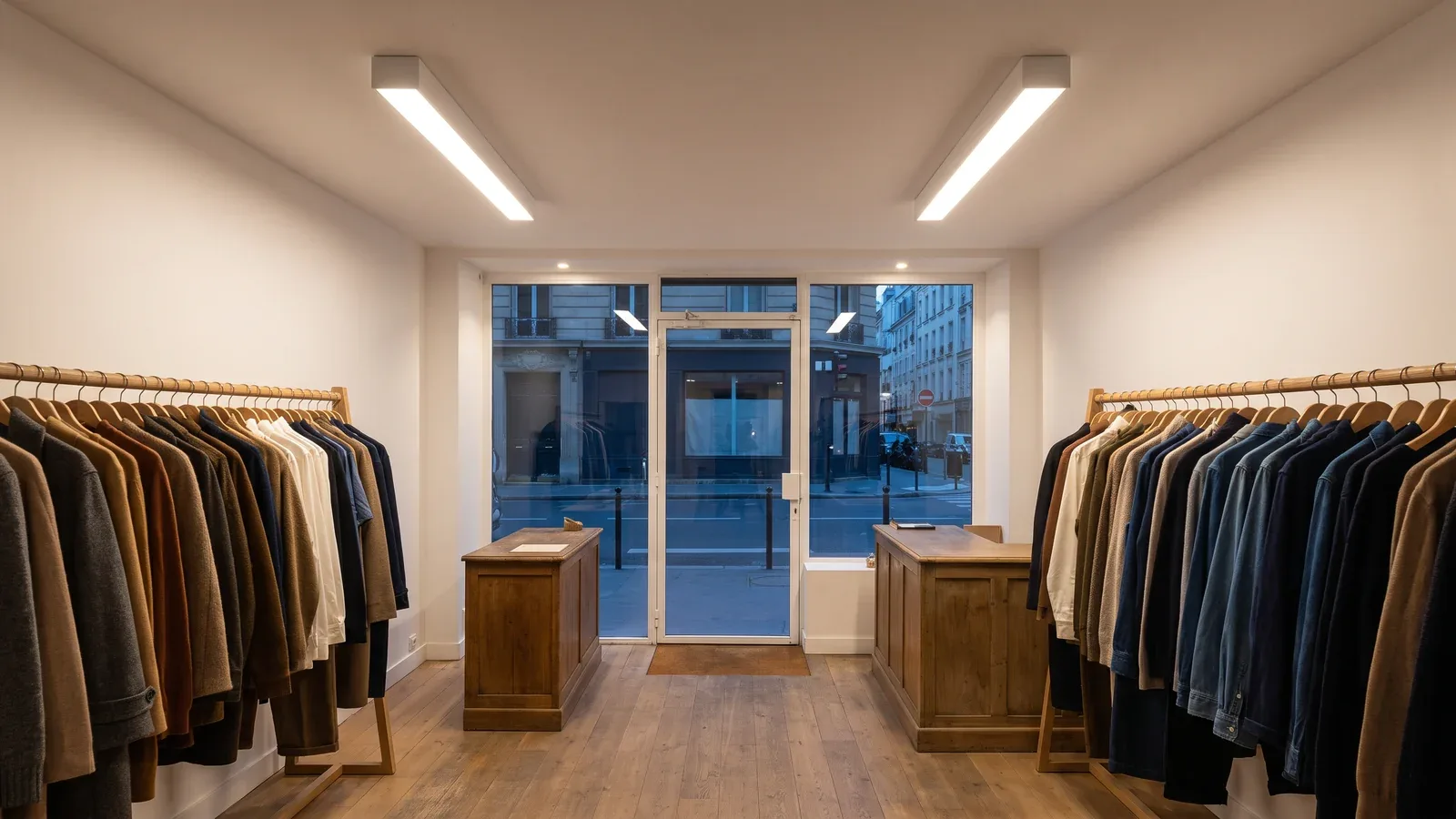 Tienda de moda con expositores de roble y líneas de luz blancas de superficie en el techo y vista hacia el escaparate al atardecer.